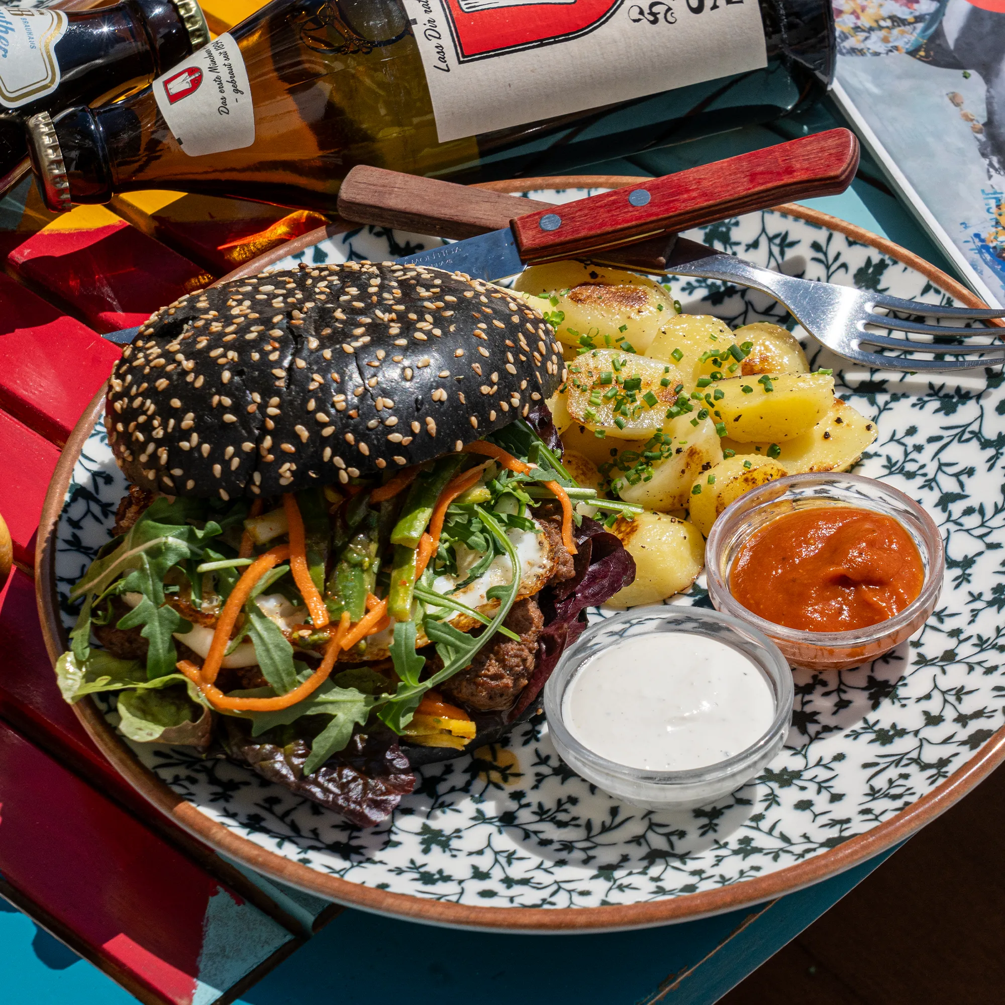 Burger im schwarzen Bun mit Bratkartoffeln und Dips auf gemustertem Teller